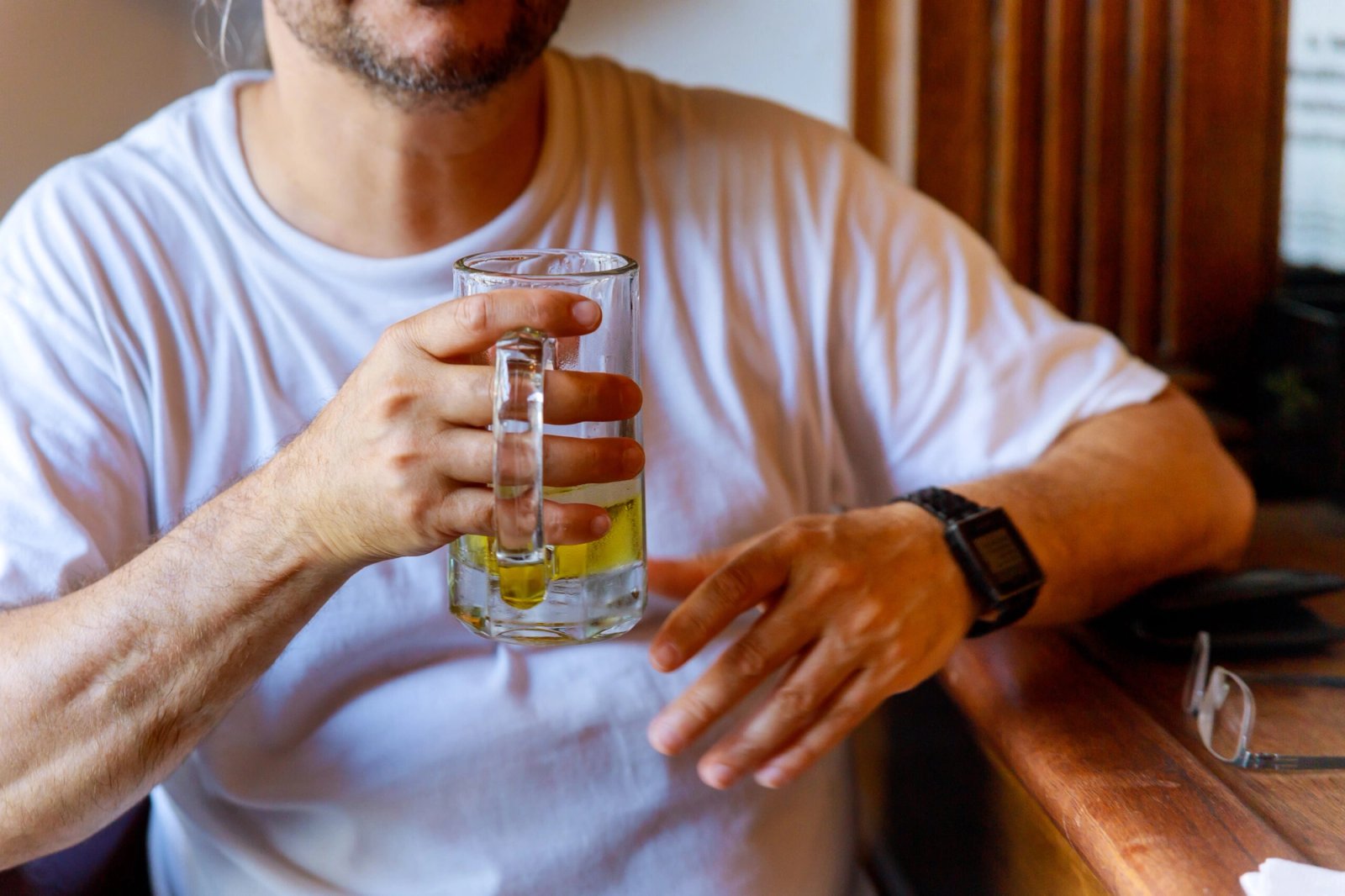 Hombre en camiseta blanca con una jarra de cerveza en la mano, sentado en la barra de un bar
