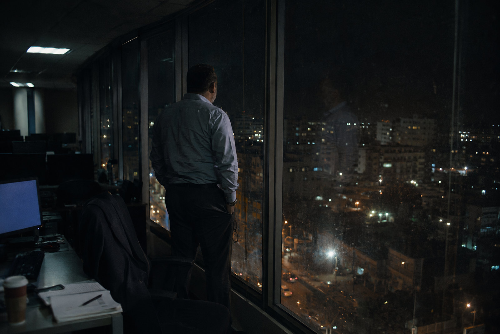 Hombre de espaldas frente al ventanal de una oficina vacía de noche, mirando las luces de la ciudad