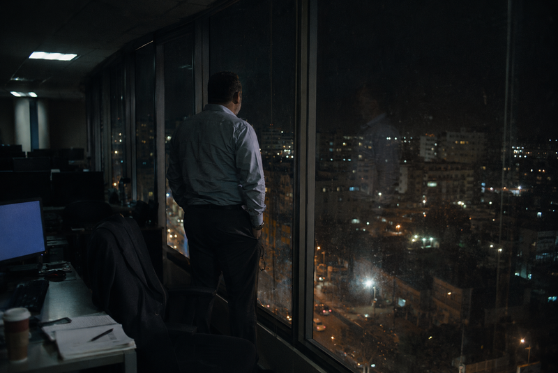 Hombre de espaldas frente al ventanal de una oficina vacía de noche, mirando las luces de la ciudad