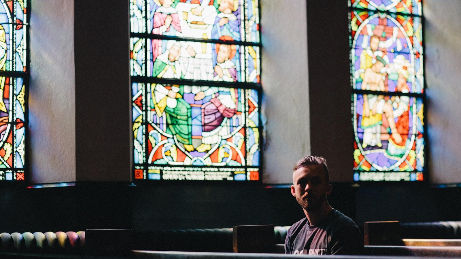 Jovem sentado num banco de igreja, com vitrais coloridos ao fundo