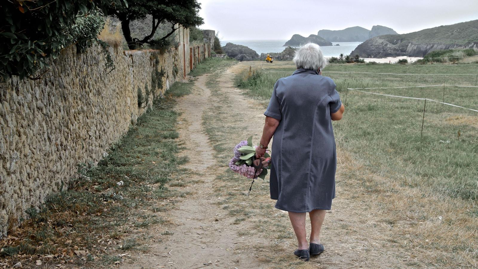 Mulher de vestido cinza caminhando por um caminho de terra junto a um muro de pedra, com flores na mão