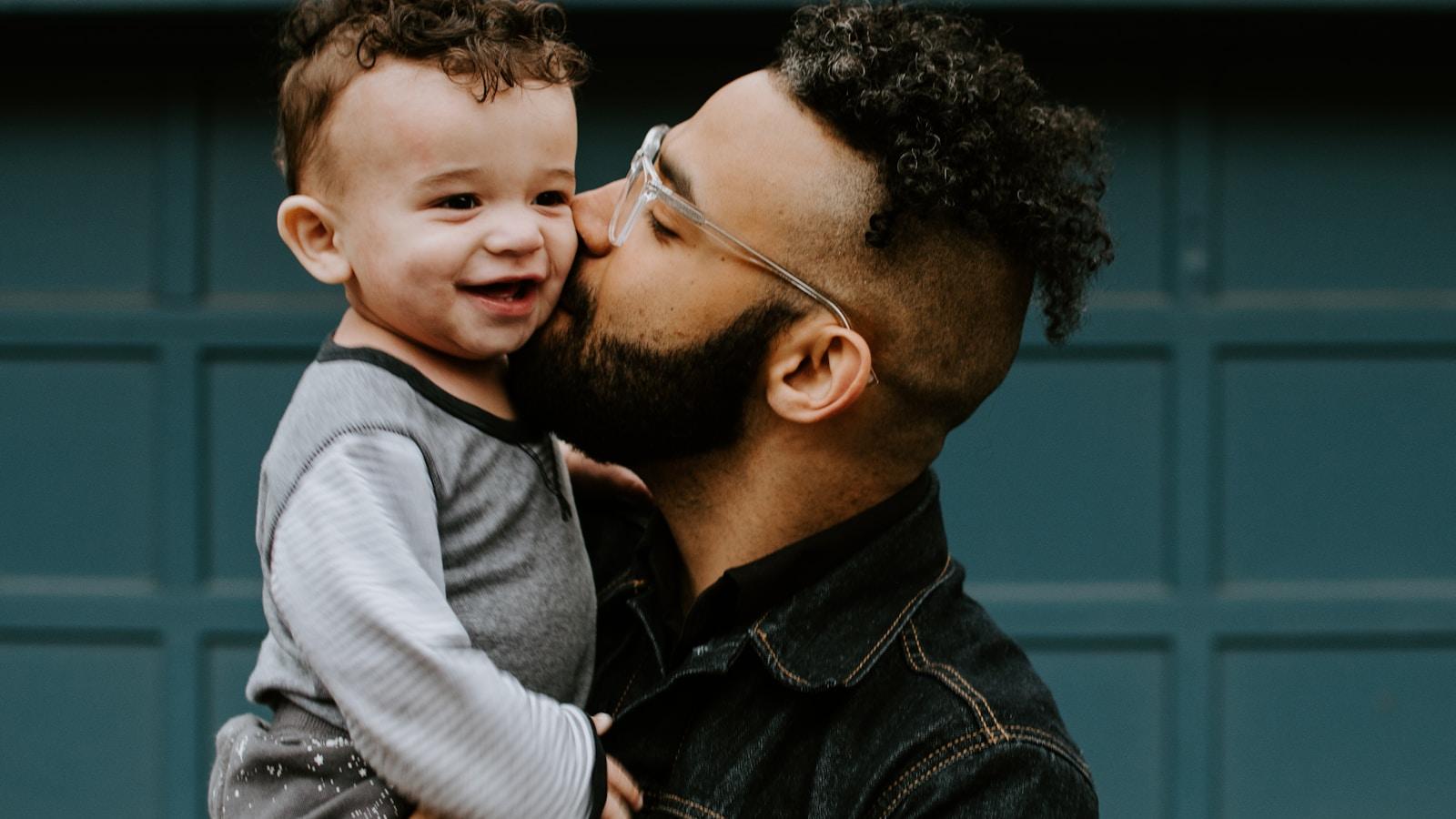 Pai beijando o rosto do filho pequeno, que sorri em seus braços