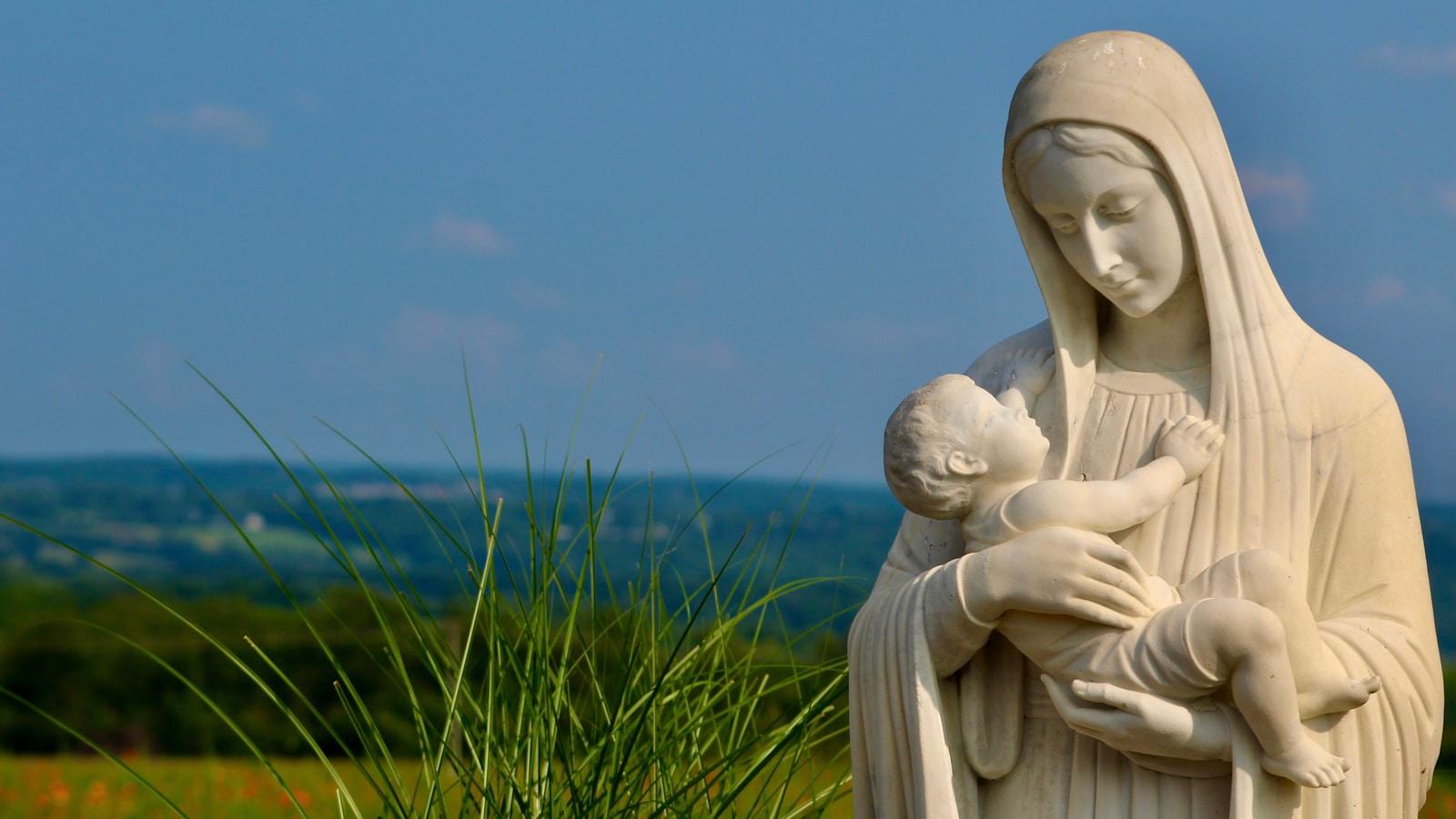 Estátua branca de Nossa Senhora com o Menino Jesus nos braços, diante de um campo