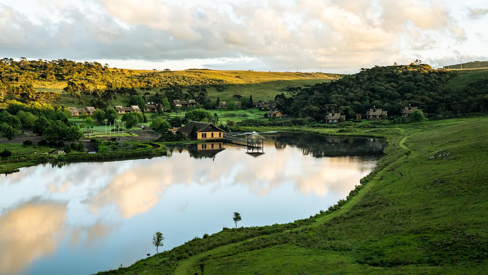 Lago cercado de colinas verdes e casas, sob céu de nuvens douradas