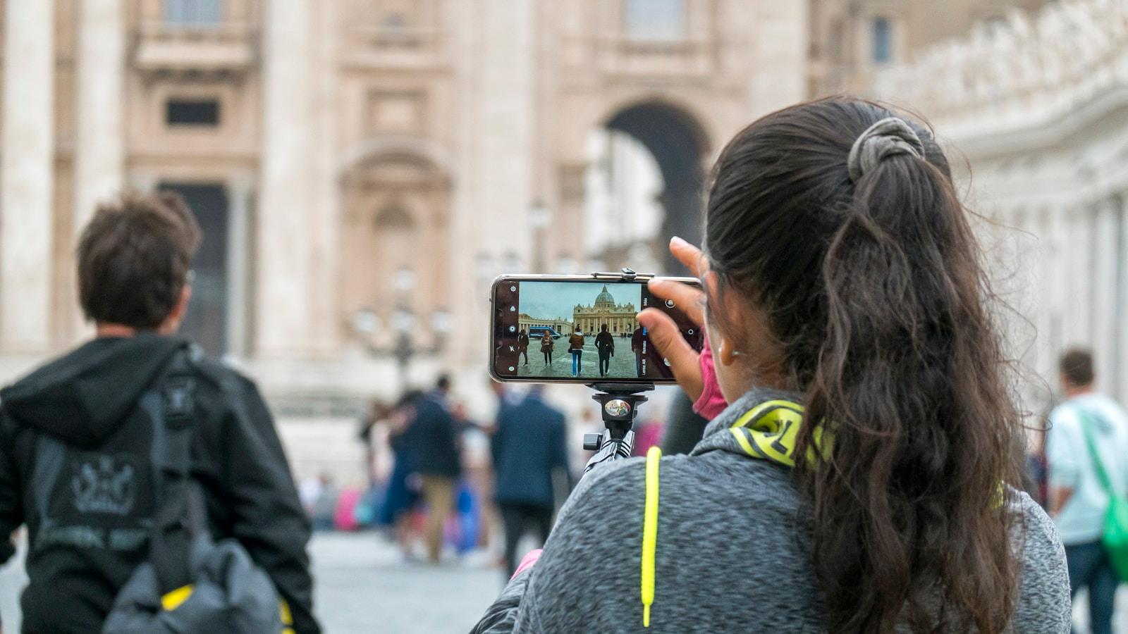 Mulher fotografando a Praça de São Pedro com o celular, vista por trás