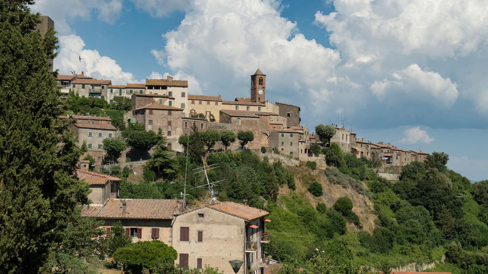 Vila italiana no alto de uma colina, com casas de pedra e torre com relógio, sob céu de nuvens