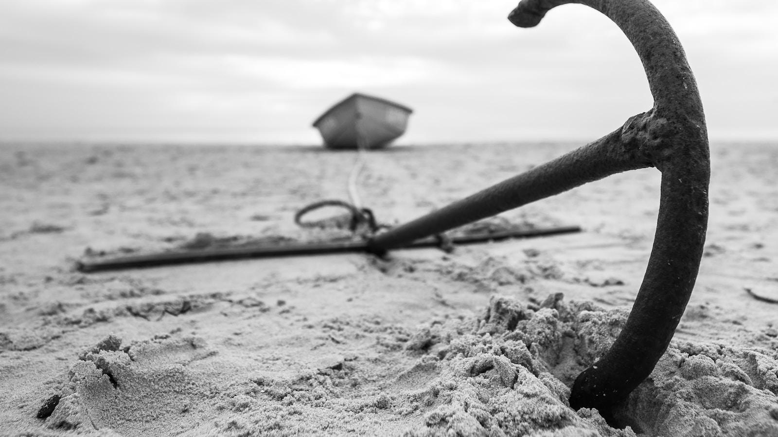 Âncora enferrujada cravada na areia da praia, com um barco ao fundo, em preto e branco