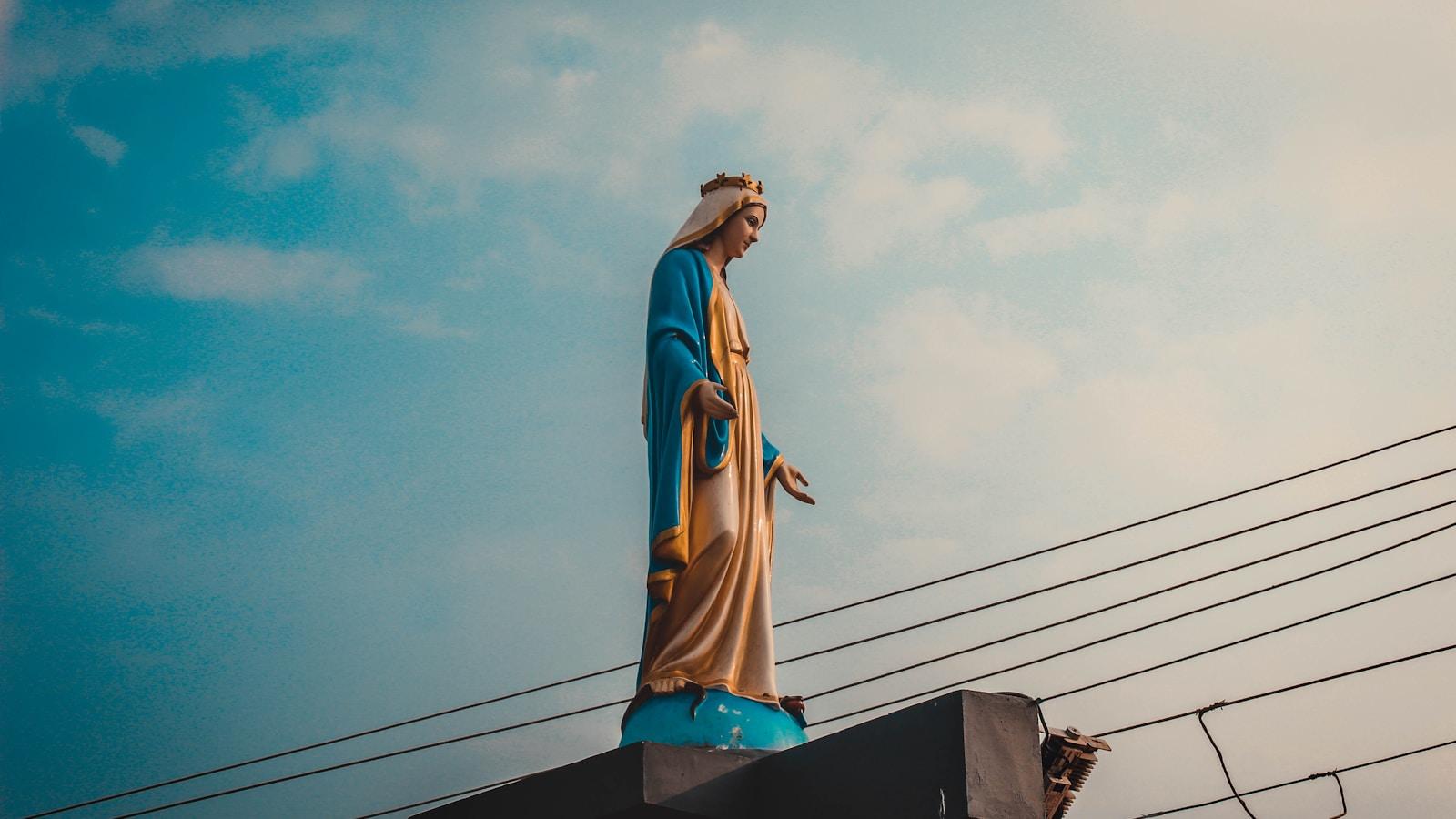 Estátua de Nossa Senhora com manto azul no alto de um telhado, contra o céu