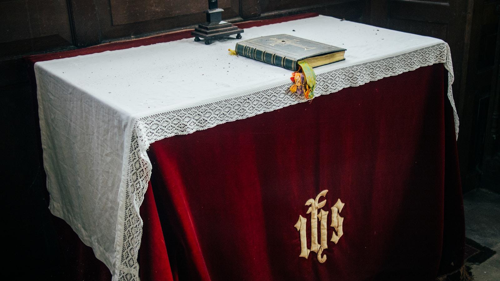 Bíblia sobre um altar coberto com pano vermelho bordado com o monograma IHS e renda branca