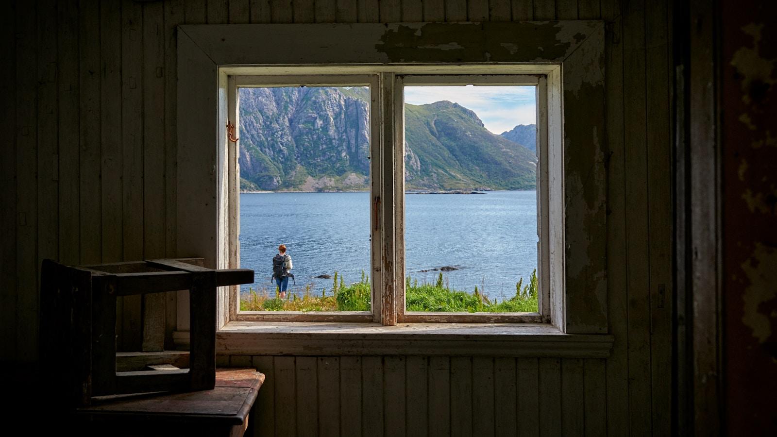 Vista pela janela de uma casa antiga para uma pessoa à beira de um lago entre montanhas