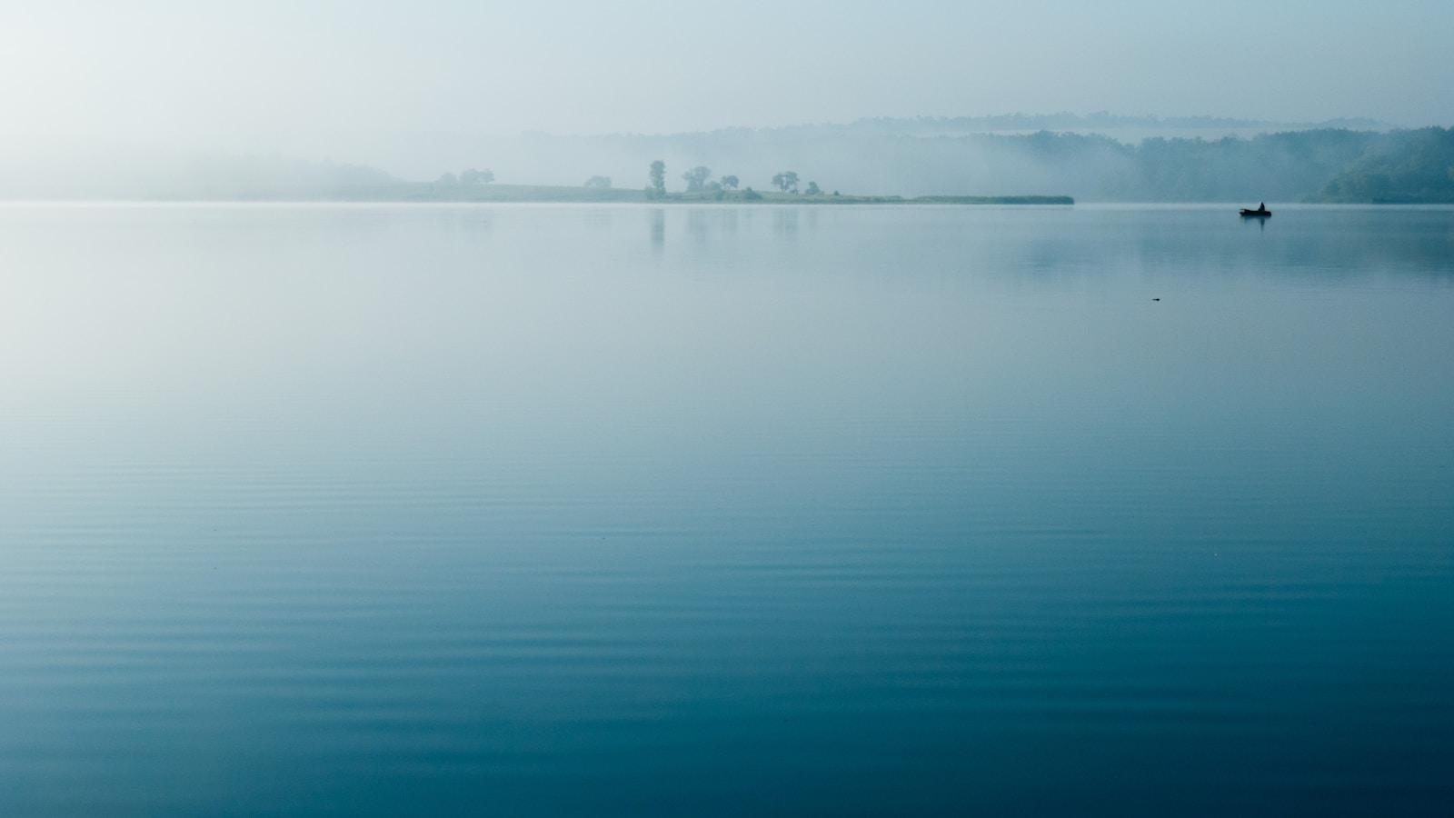 Lago de águas calmas e azuladas sob névoa, com um pequeno barco ao longe