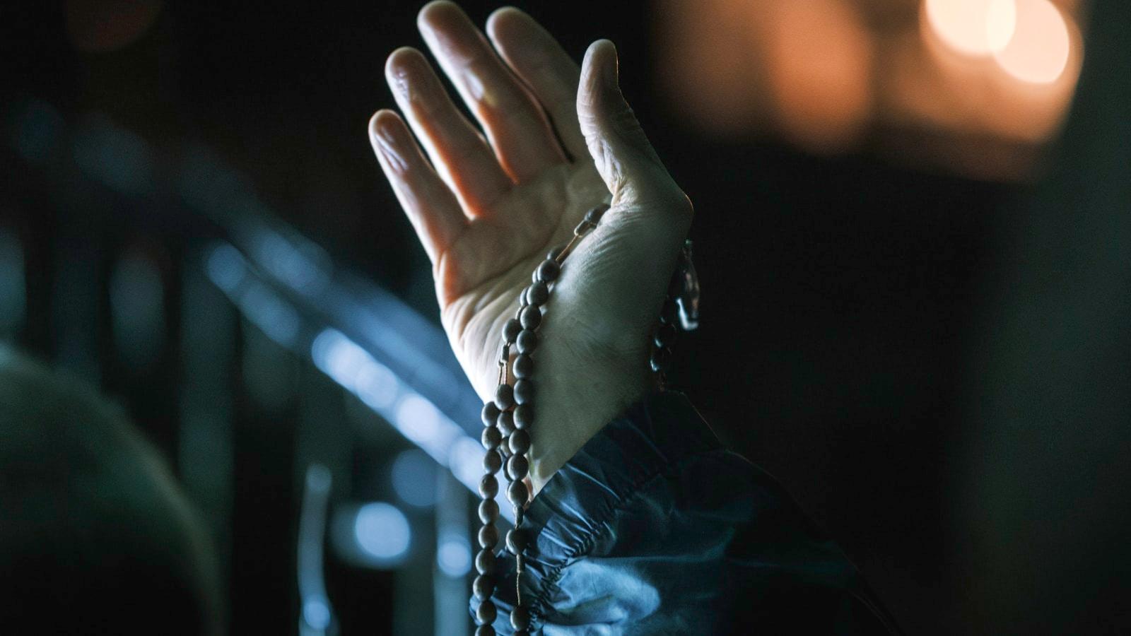 Mão erguida segurando um rosário de contas escuras, em luz baixa