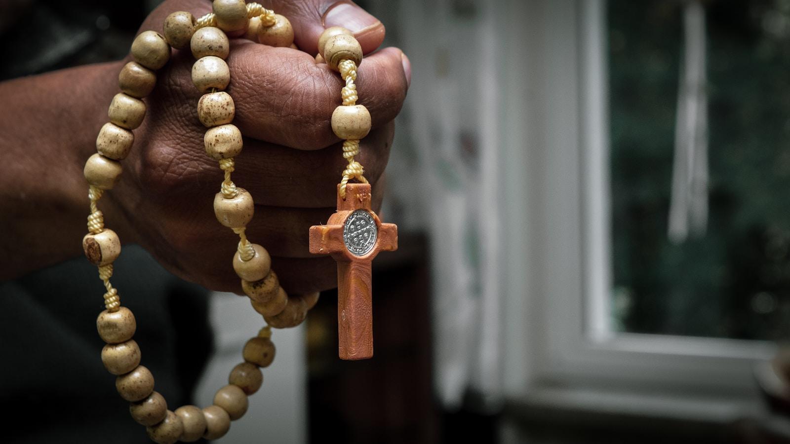 Mãos segurando um rosário de madeira com cruz de São Bento, junto a uma janela