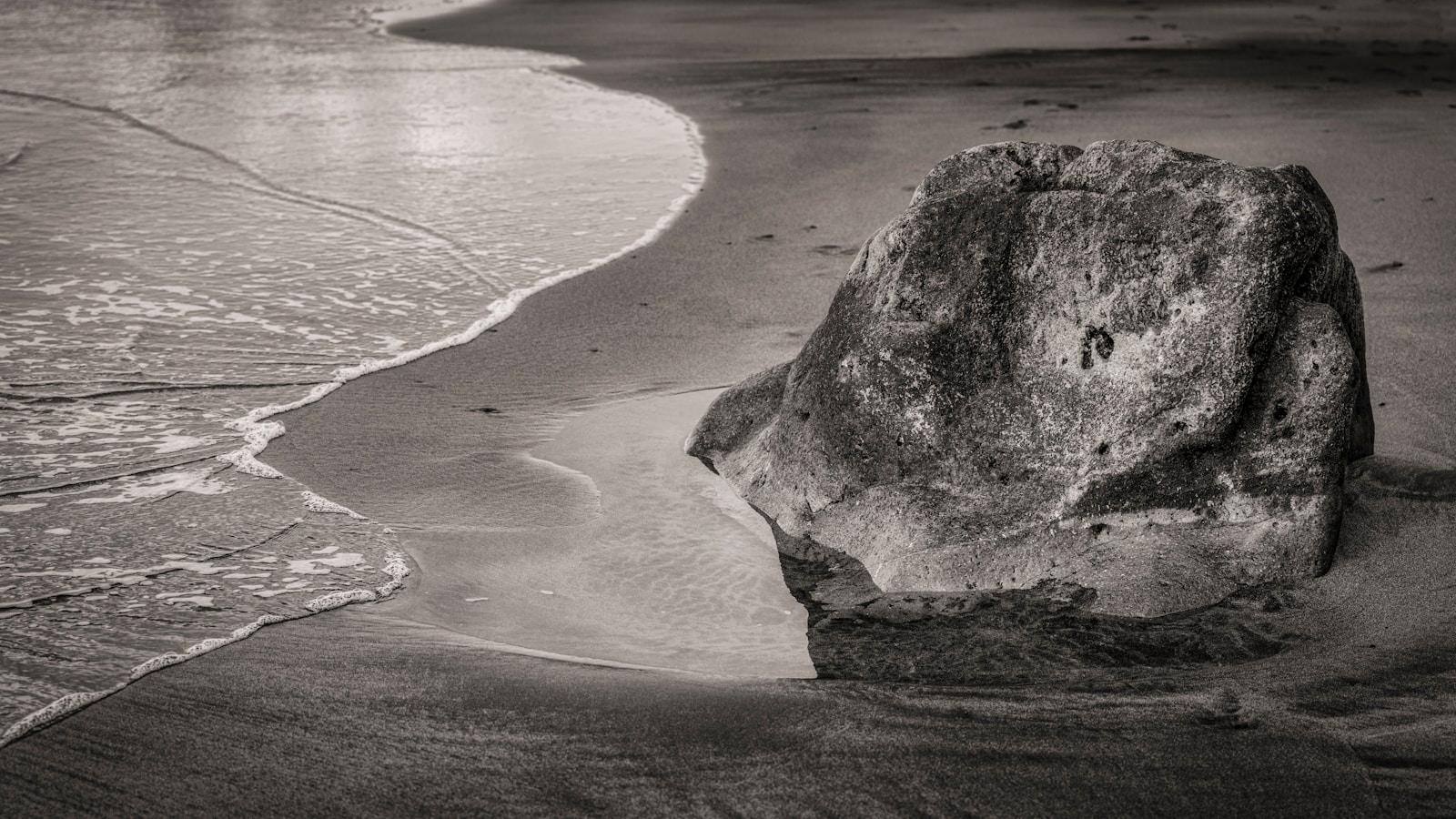 Grande rocha na areia da praia com a espuma das ondas ao lado, em preto e branco