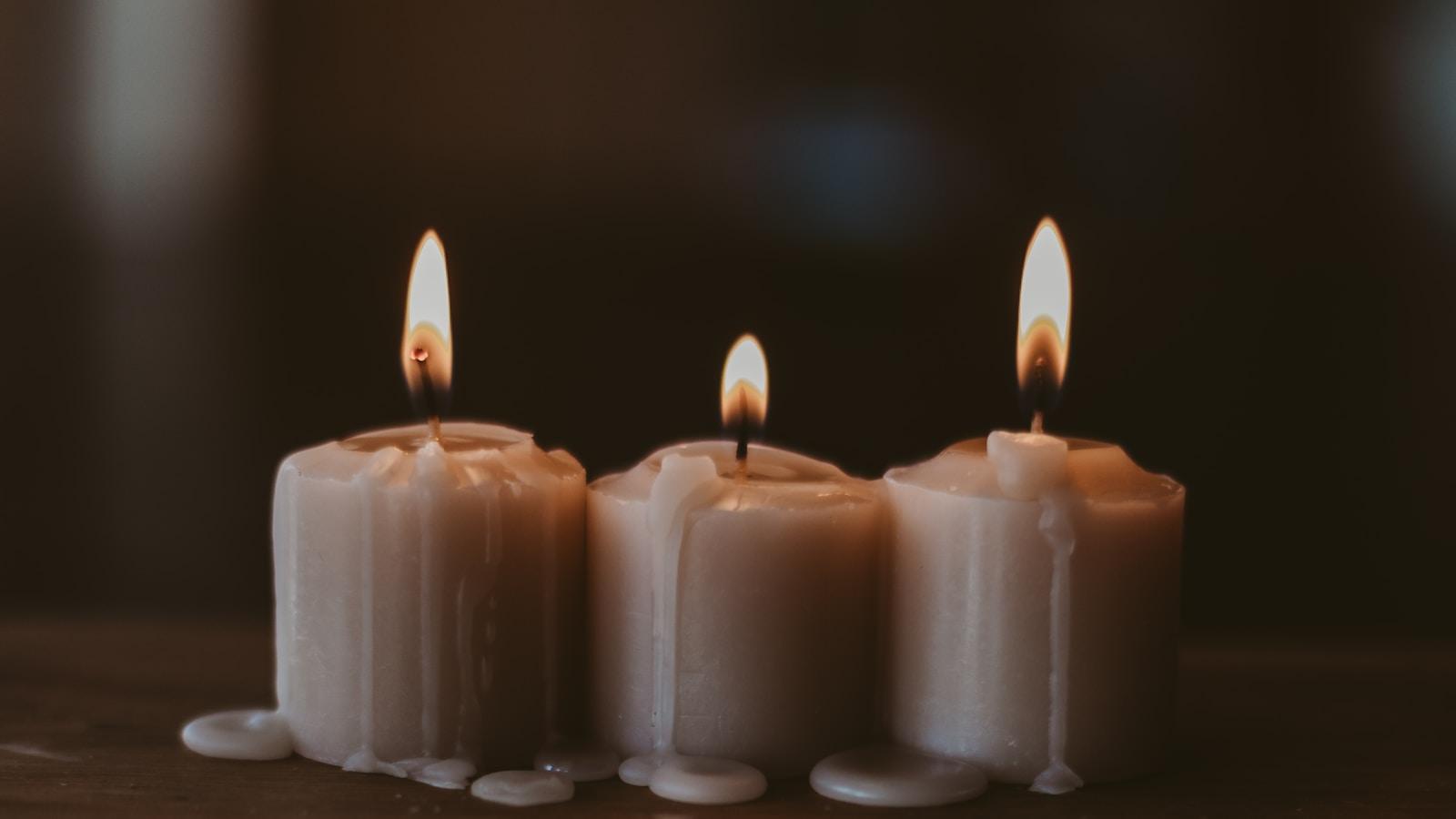 Três velas brancas acesas, lado a lado, sobre uma mesa de madeira