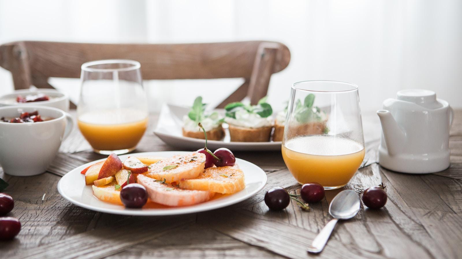 Mesa de café da manhã com frutas, suco e pão, junto a uma janela clara