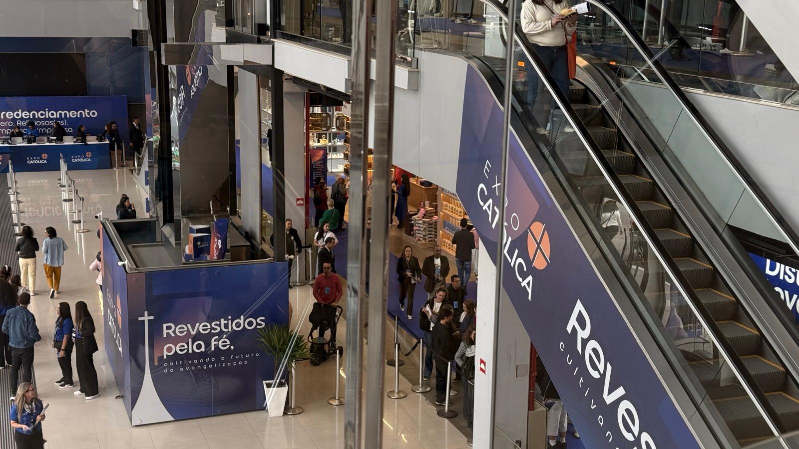 Saguão da ExpoCatólica com escadas rolantes, visitantes e painéis azuis do evento com o lema "Revestidos pela fé"