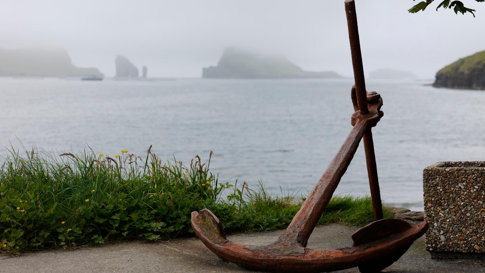 Âncora antiga de madeira e ferro à beira-mar, com rochedos na névoa ao fundo