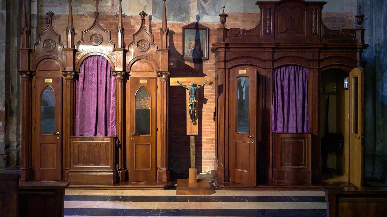 Confessionários de madeira com cortinas roxas e um crucifixo, no interior de uma igreja