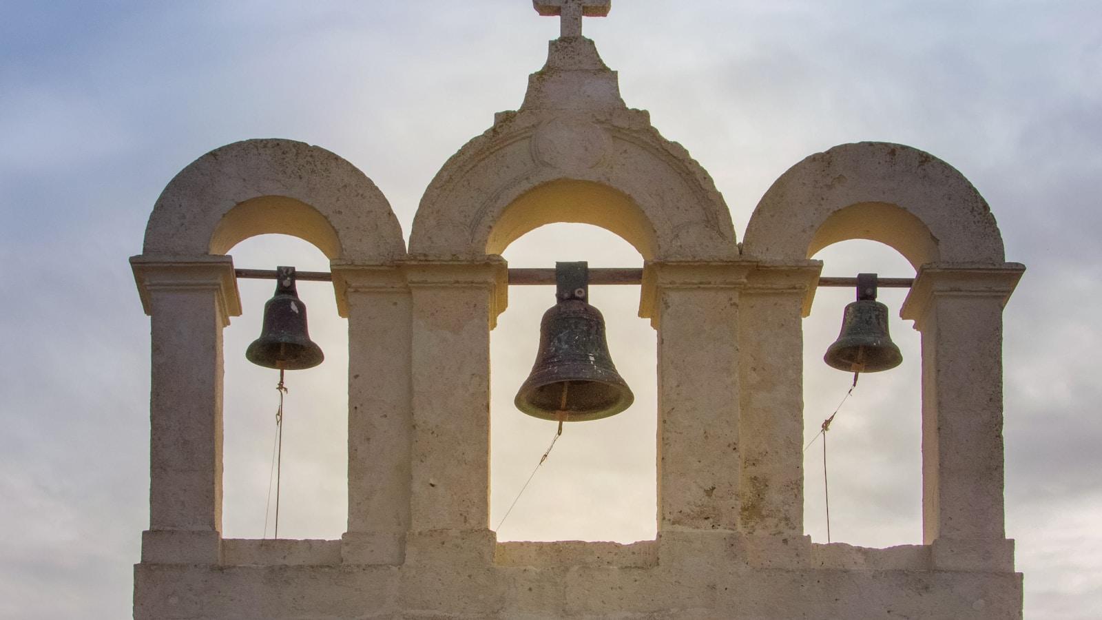 Campanário de pedra com três sinos sob arcos, contra o céu nublado