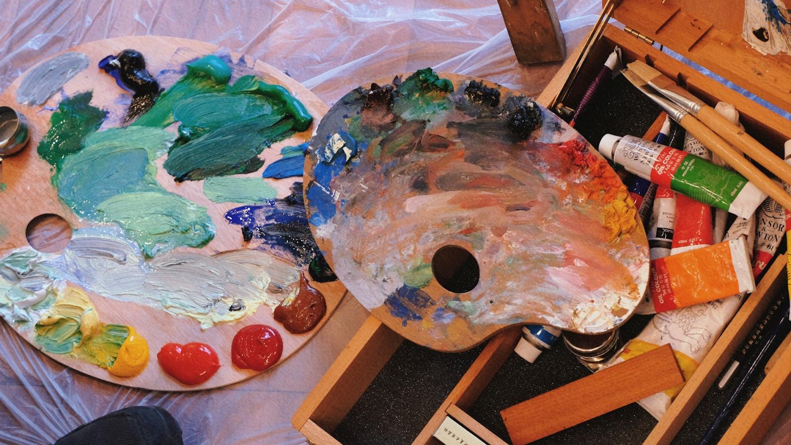 Duas paletas de pintor cobertas de tintas coloridas, com tubos de tinta ao lado