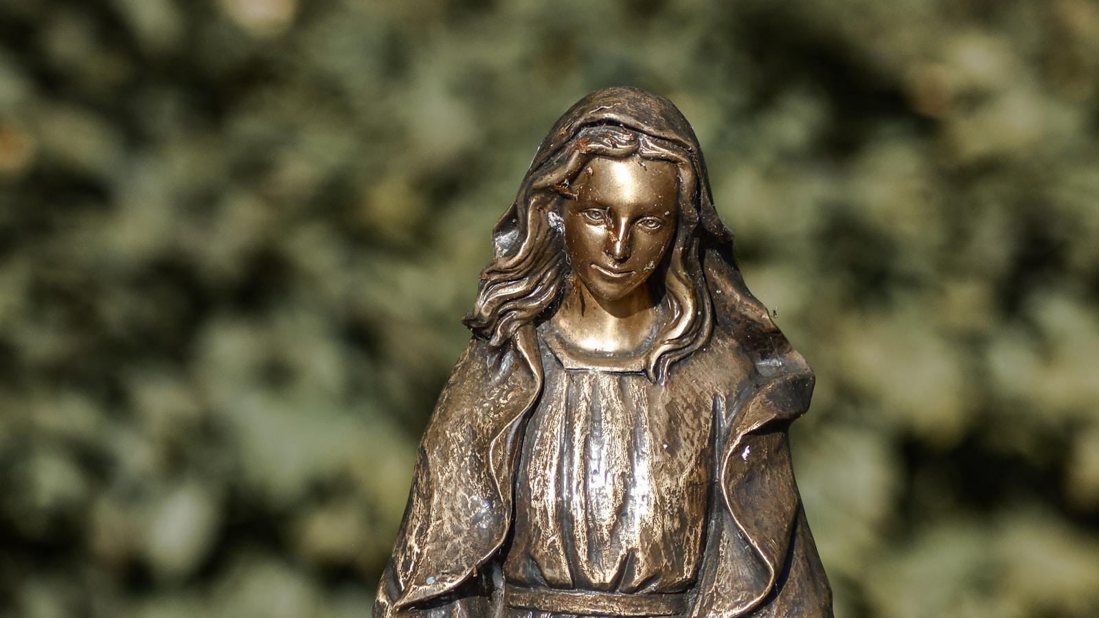 Estátua de bronze de Nossa Senhora com leve sorriso, diante de folhagem verde