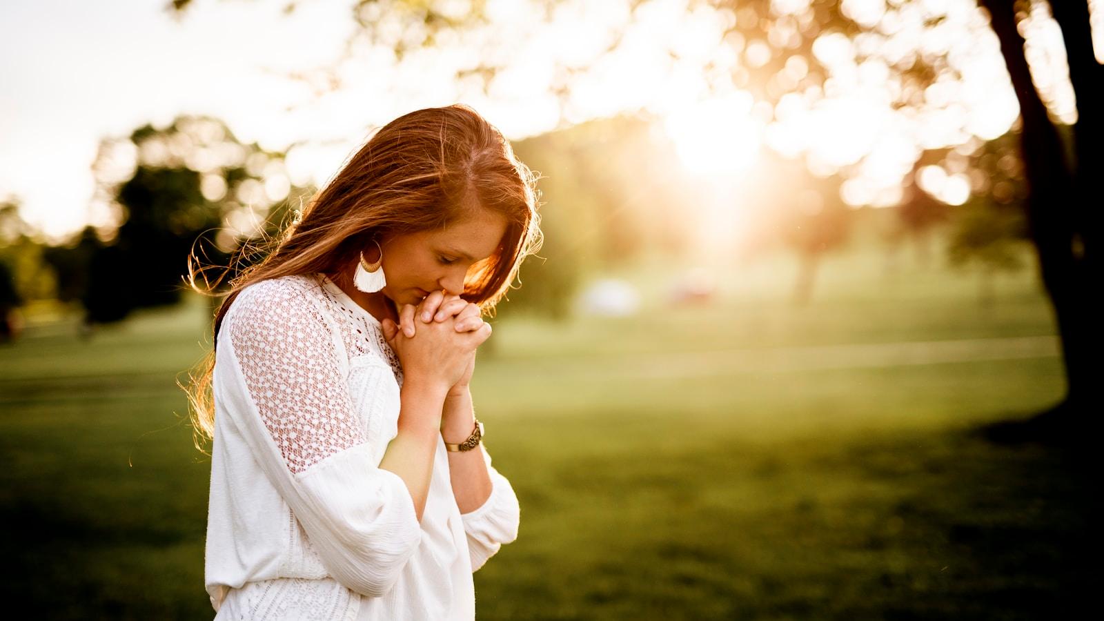 Jovem de blusa branca com as mãos juntas em oração, num parque ao pôr do sol