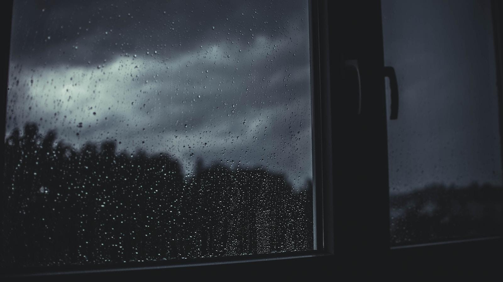 Gotas de chuva numa janela, com nuvens escuras e árvores ao fundo