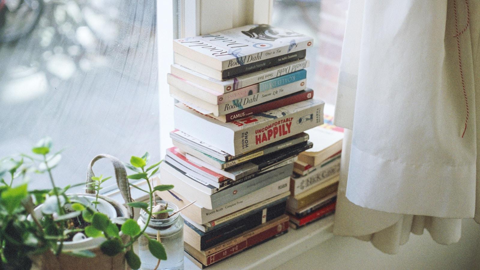 Pilha de livros junto a uma janela com cortina branca e uma planta