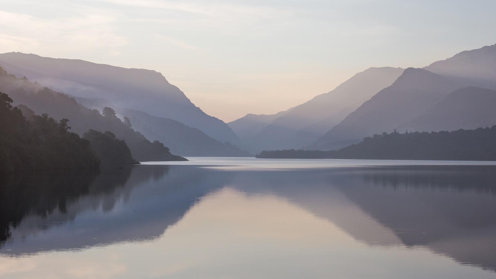 Lago calmo entre montanhas, com névoa ao amanhecer