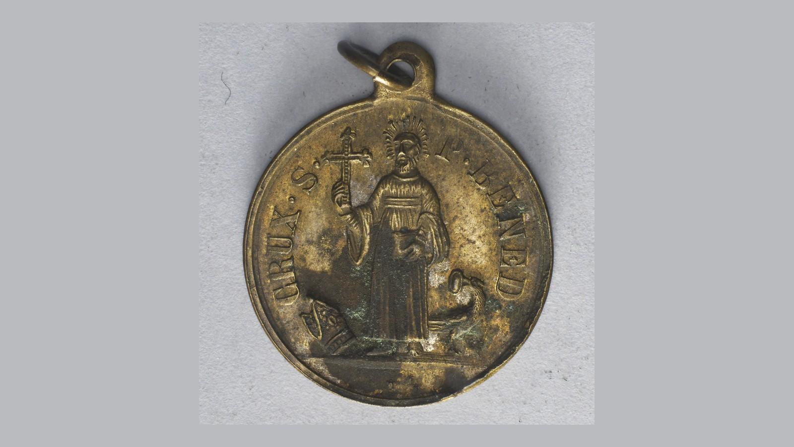 Frente de uma antiga Medalha de São Bento em latão: o santo com a cruz e o livro, a mitra aos pés e o cálice com a serpente, sob a inscrição CRUX S. P. BENED