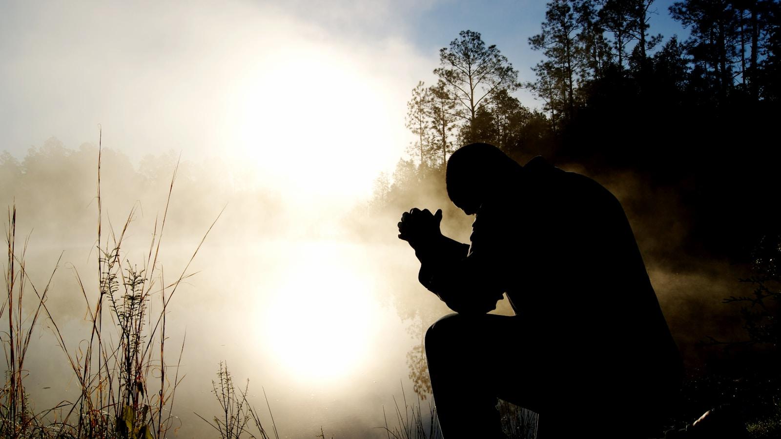 Silhueta de um homem ajoelhado em oração diante de um lago com névoa, ao nascer do sol
