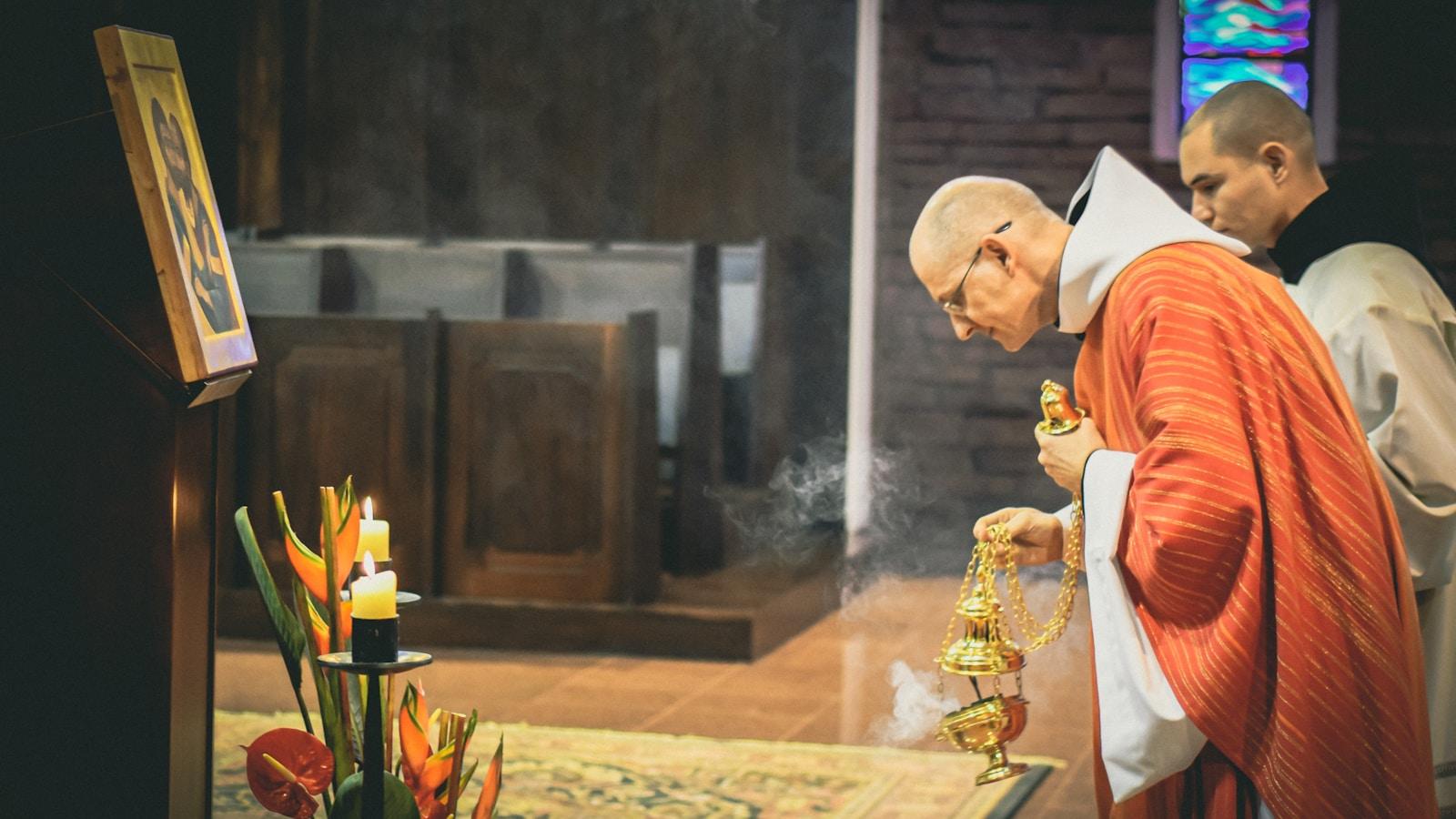 Sacerdote de casula vermelha incensando o altar, com um monge ao lado, numa capela
