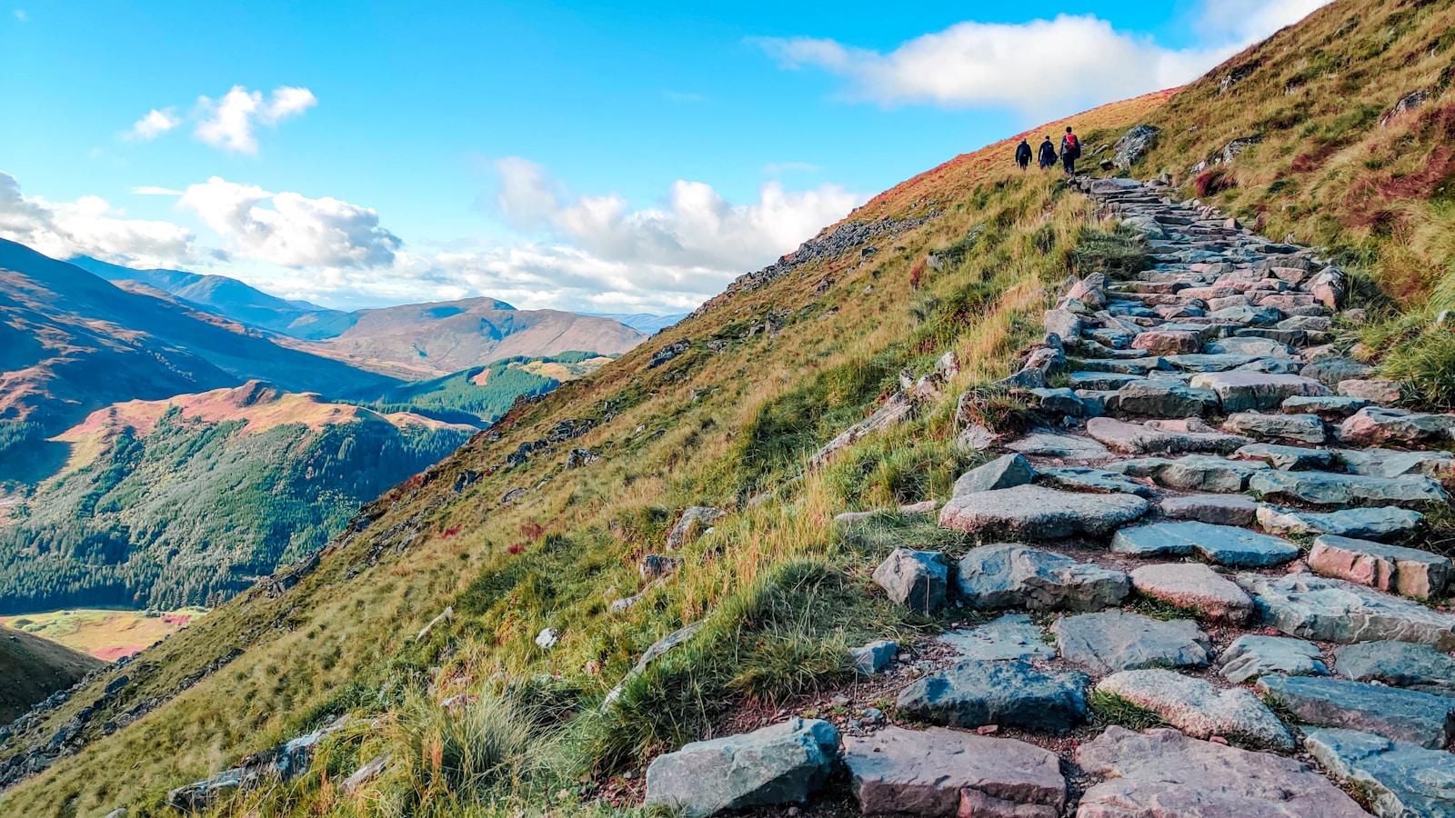 Trilha de pedras subindo uma montanha, com caminhantes ao longe e vale ao fundo