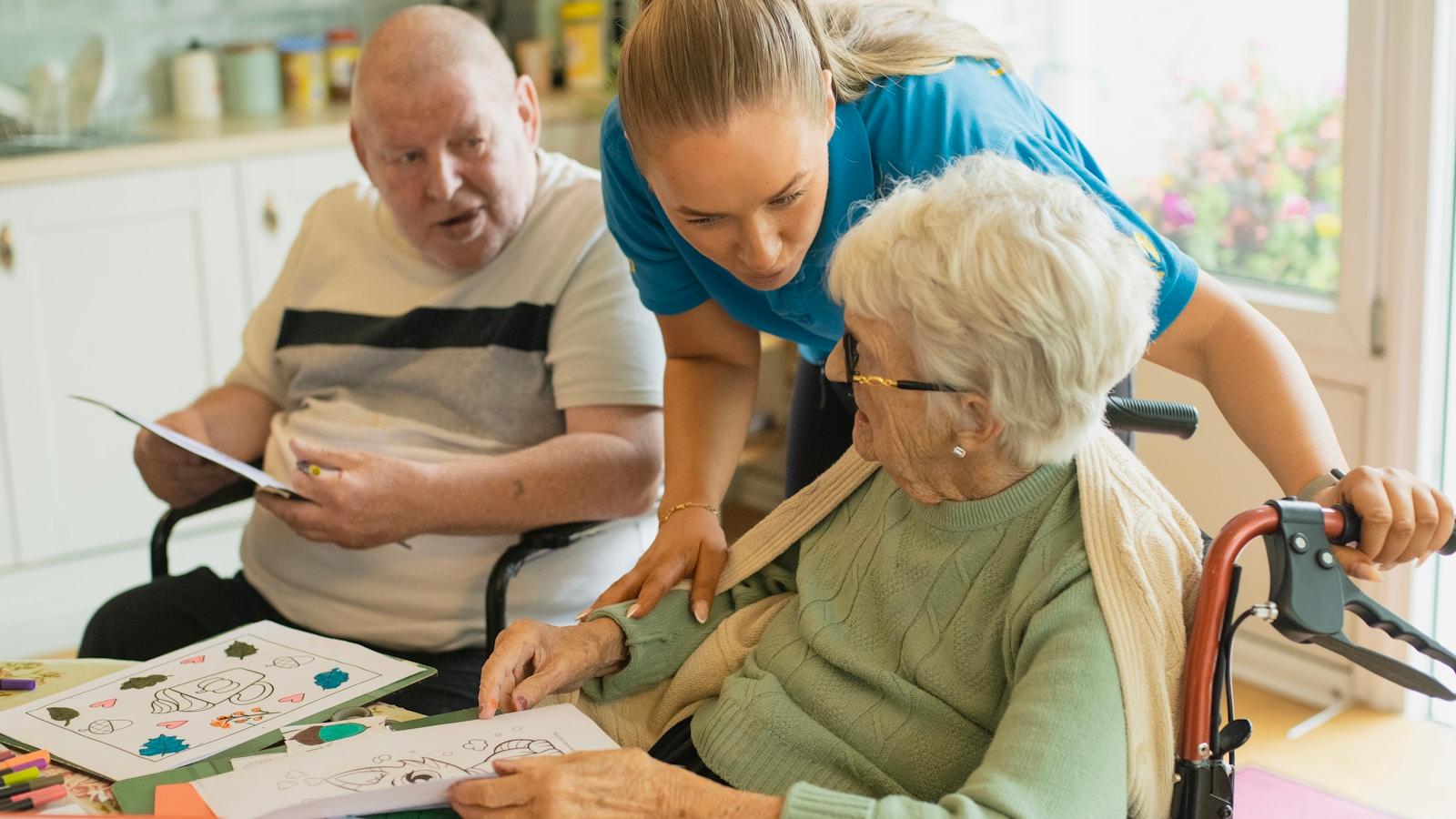 Cuidadora atendendo uma senhora idosa em cadeira de rodas, com um senhor ao lado