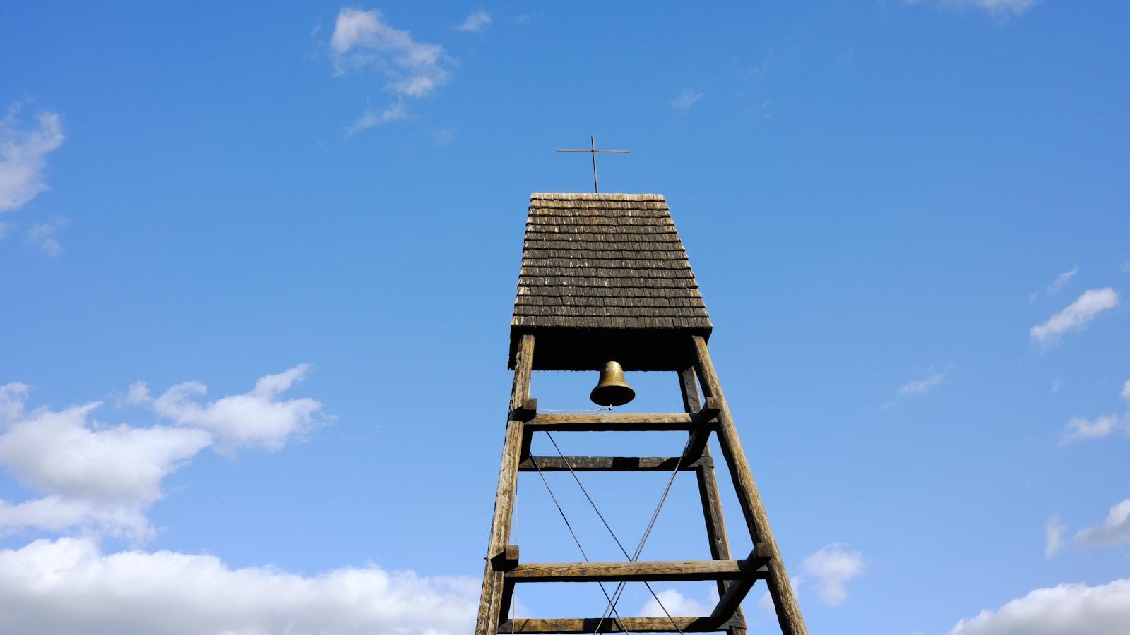 Torre de madeira com sino e uma cruz no topo, contra o céu azul