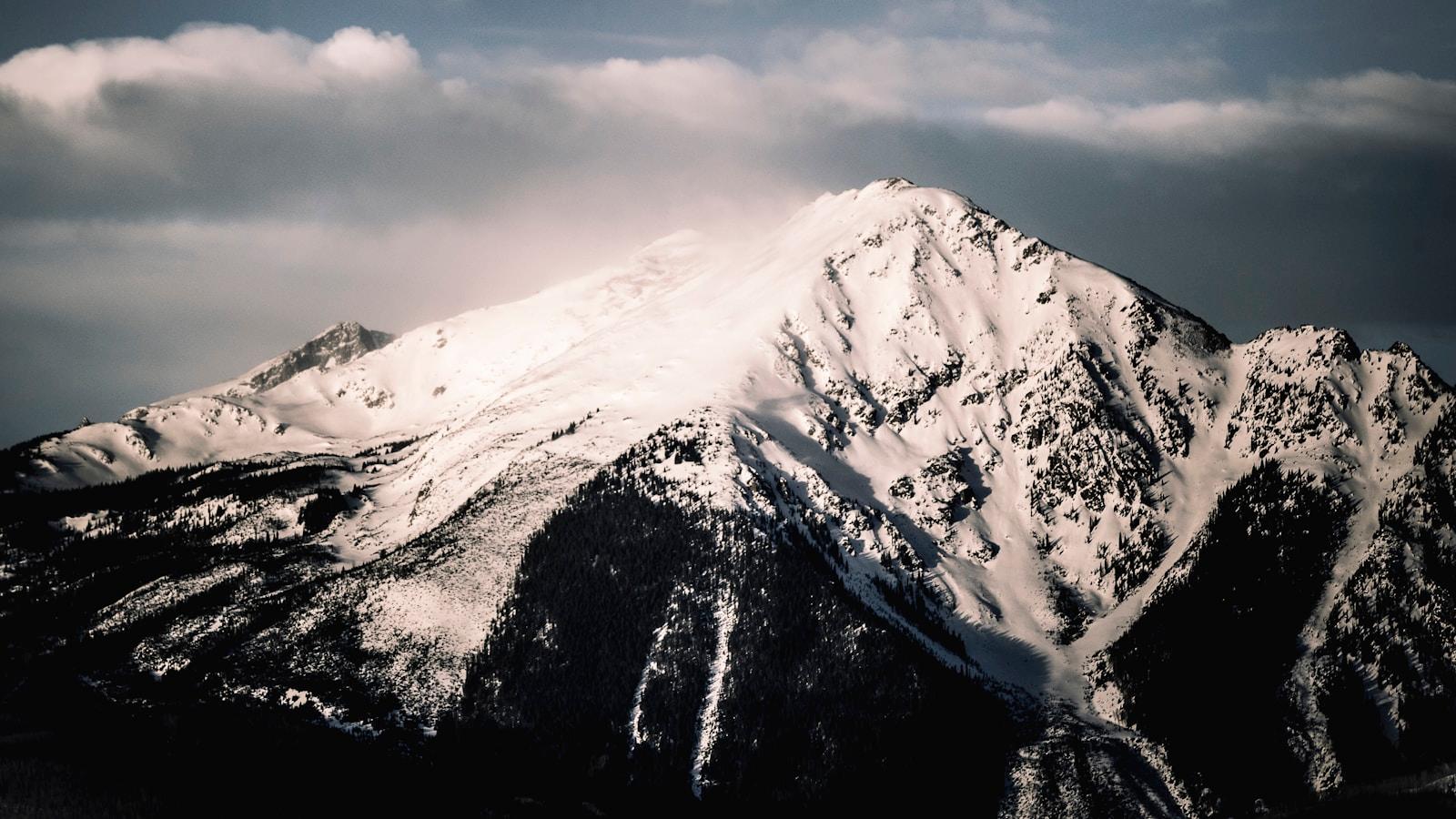 Pico de montanha coberto de neve, com a luz atravessando as nuvens no topo