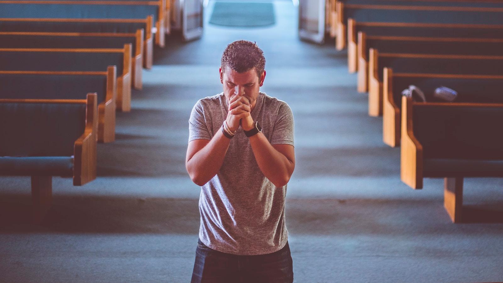 Homem de pé no corredor de uma igreja, entre os bancos, com as mãos juntas em oração
