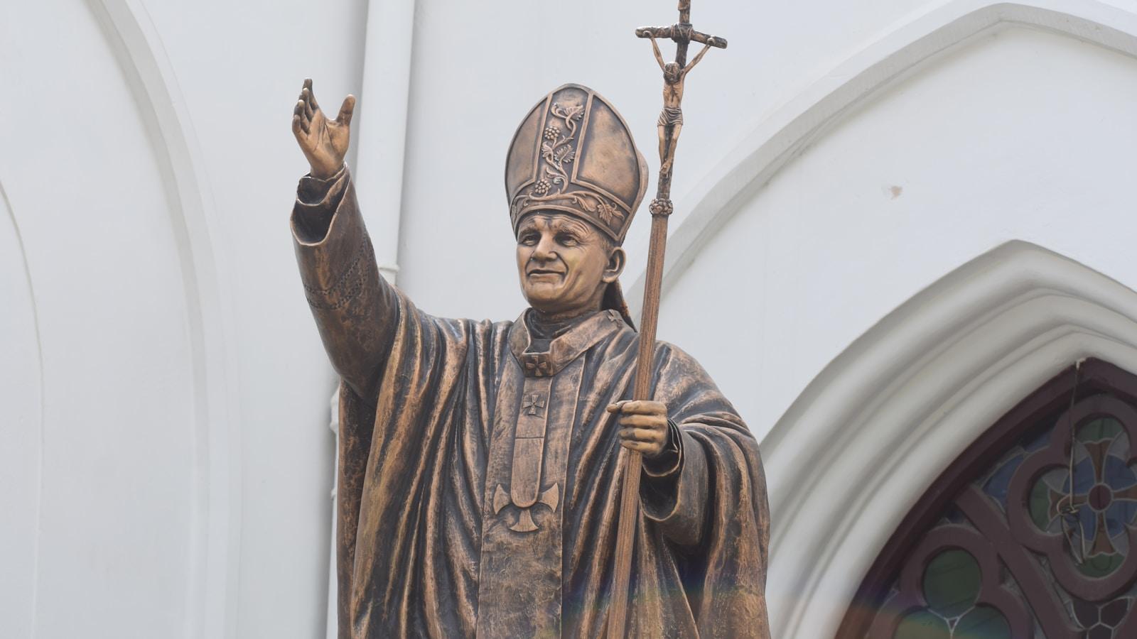 Estátua de bronze de São João Paulo II com a mão erguida e o báculo, diante de uma igreja