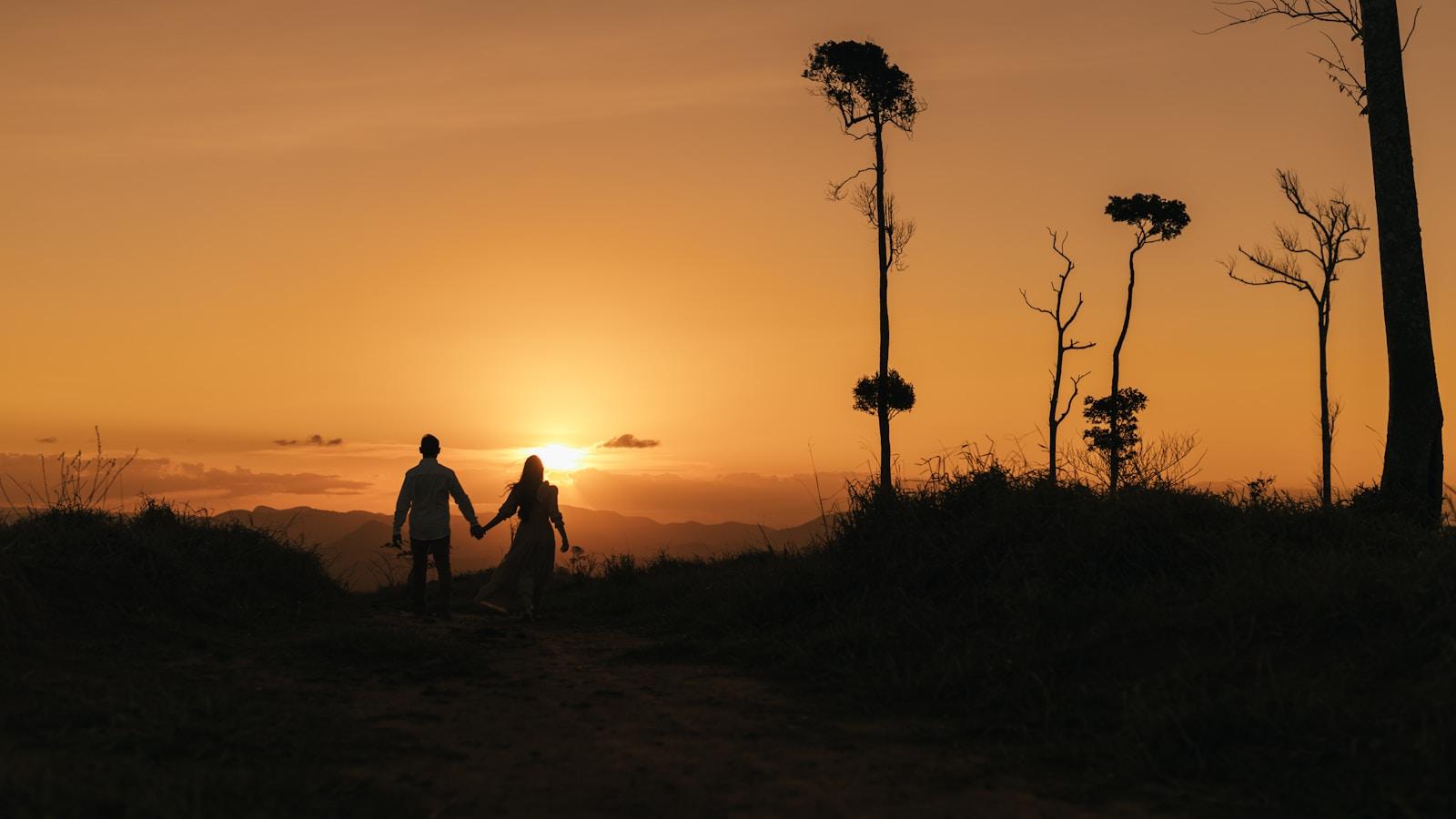 Duas pessoas de mãos dadas caminhando por uma trilha ao pôr do sol