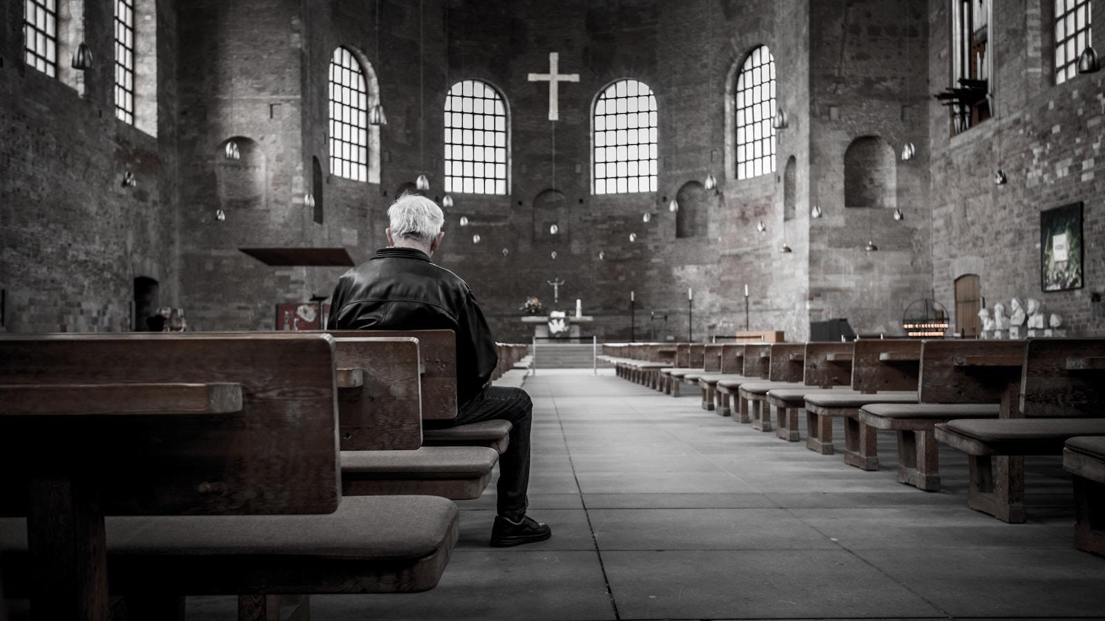 Homem sentado sozinho num banco de igreja, diante de uma cruz e janelas em arco