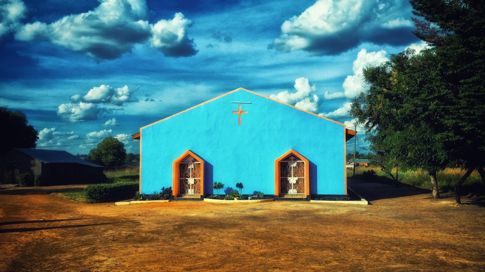 Pequena igreja de paredes azuis com uma cruz na fachada, em terreno de terra vermelha, sob céu de nuvens