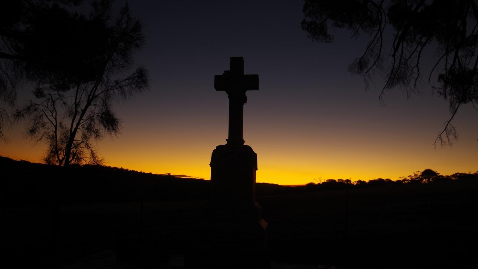 Silhueta de uma cruz de pedra contra o céu do anoitecer, entre árvores