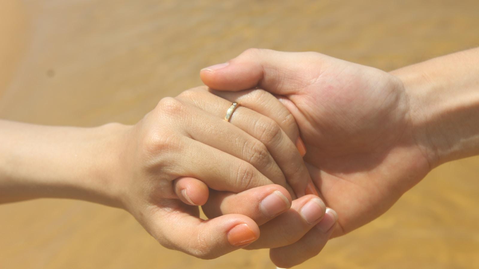 Duas mãos entrelaçadas, uma delas com aliança, sobre fundo de areia