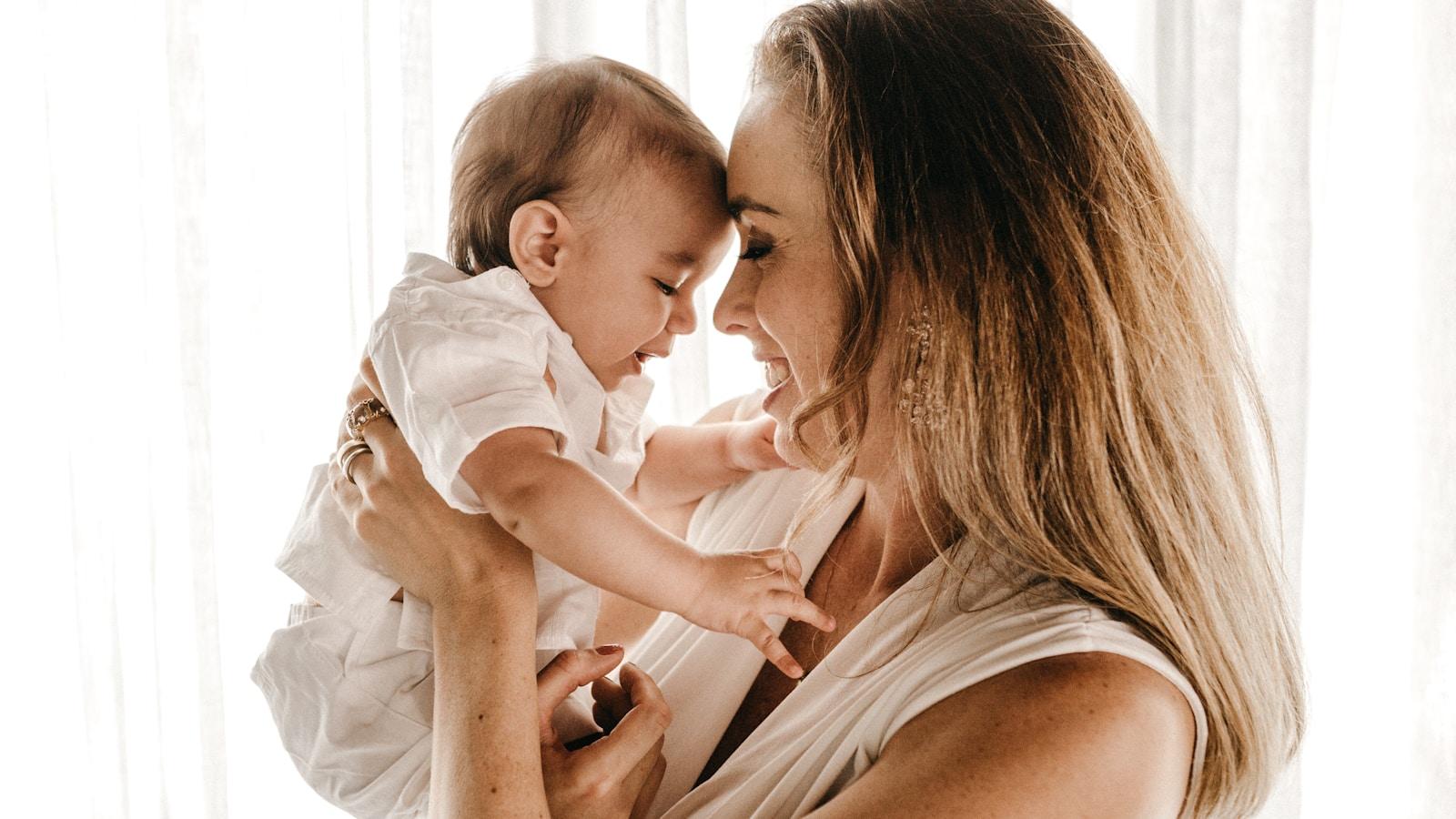 Mãe sorrindo com o bebê no colo, testa com testa, diante de uma janela clara