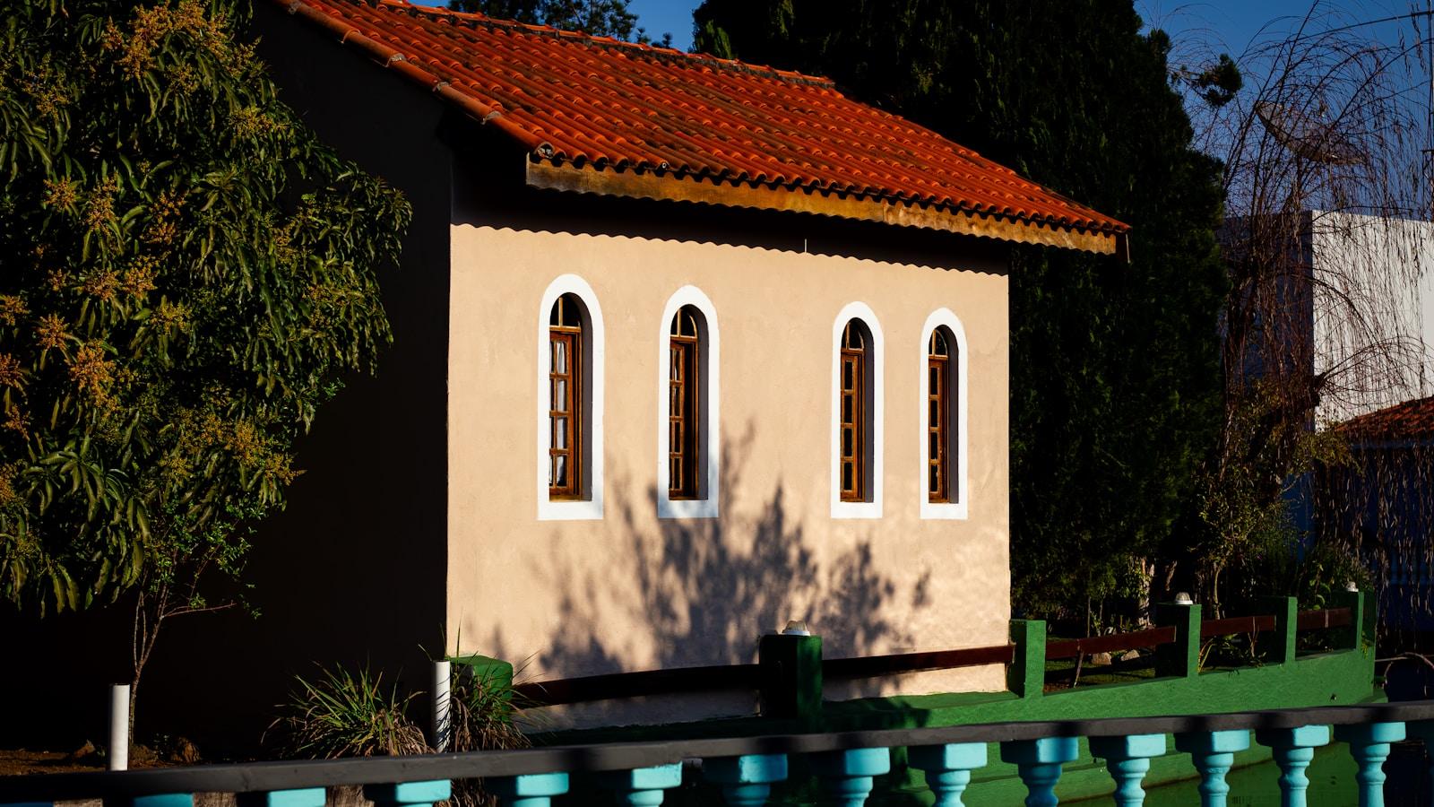 Pequena capela de paredes bege e telhado de telhas vermelhas, com janelas em arco, entre árvores