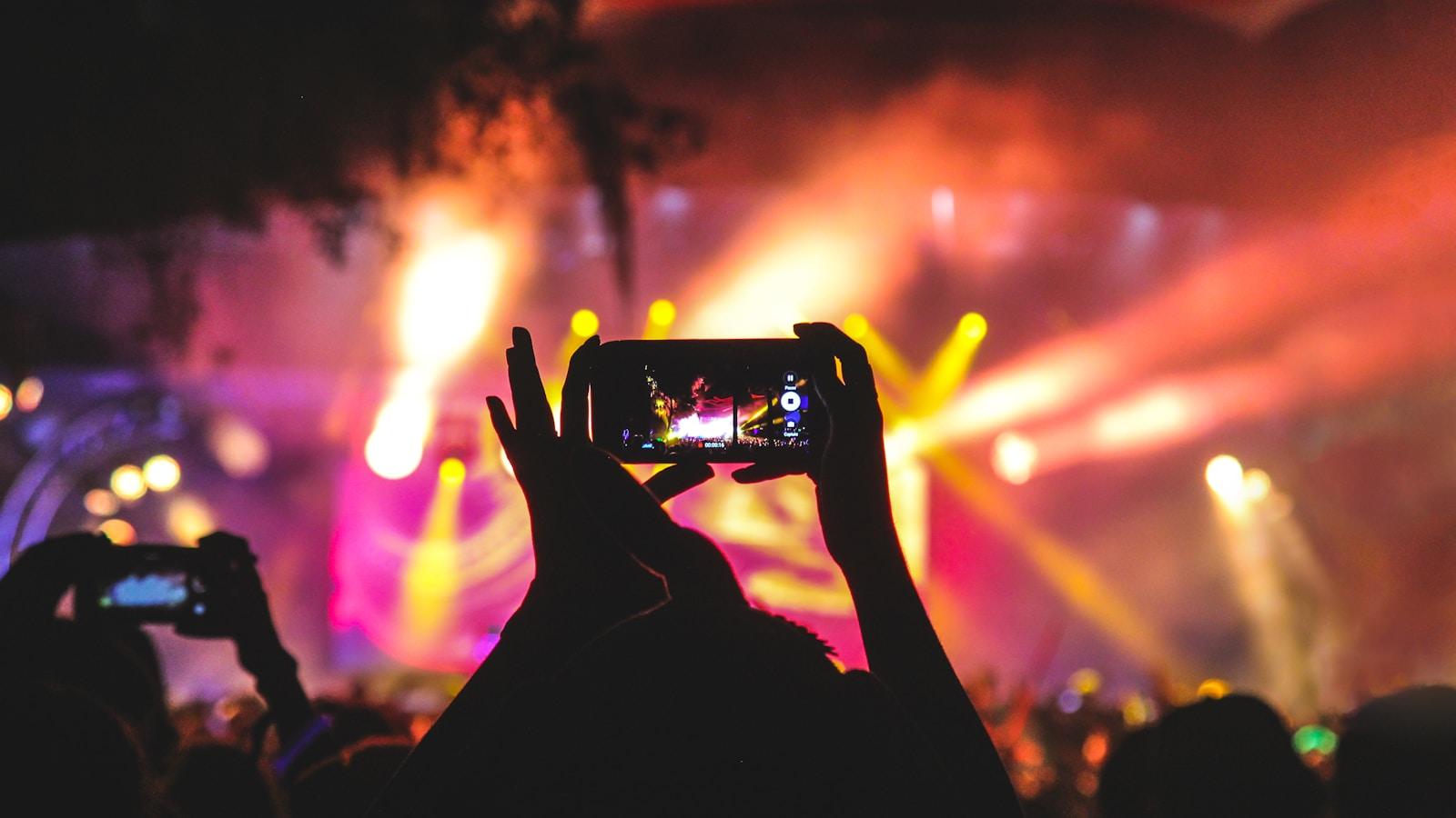 Pessoas filmando um show com celulares erguidos, sob luzes coloridas