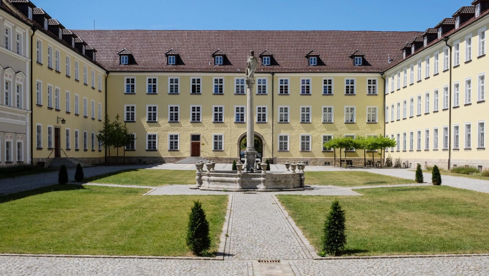 Pátio interno da abadia beneditina de Metten, na Baviera: alas de fachada amarela em volta de um jardim com fonte de pedra.