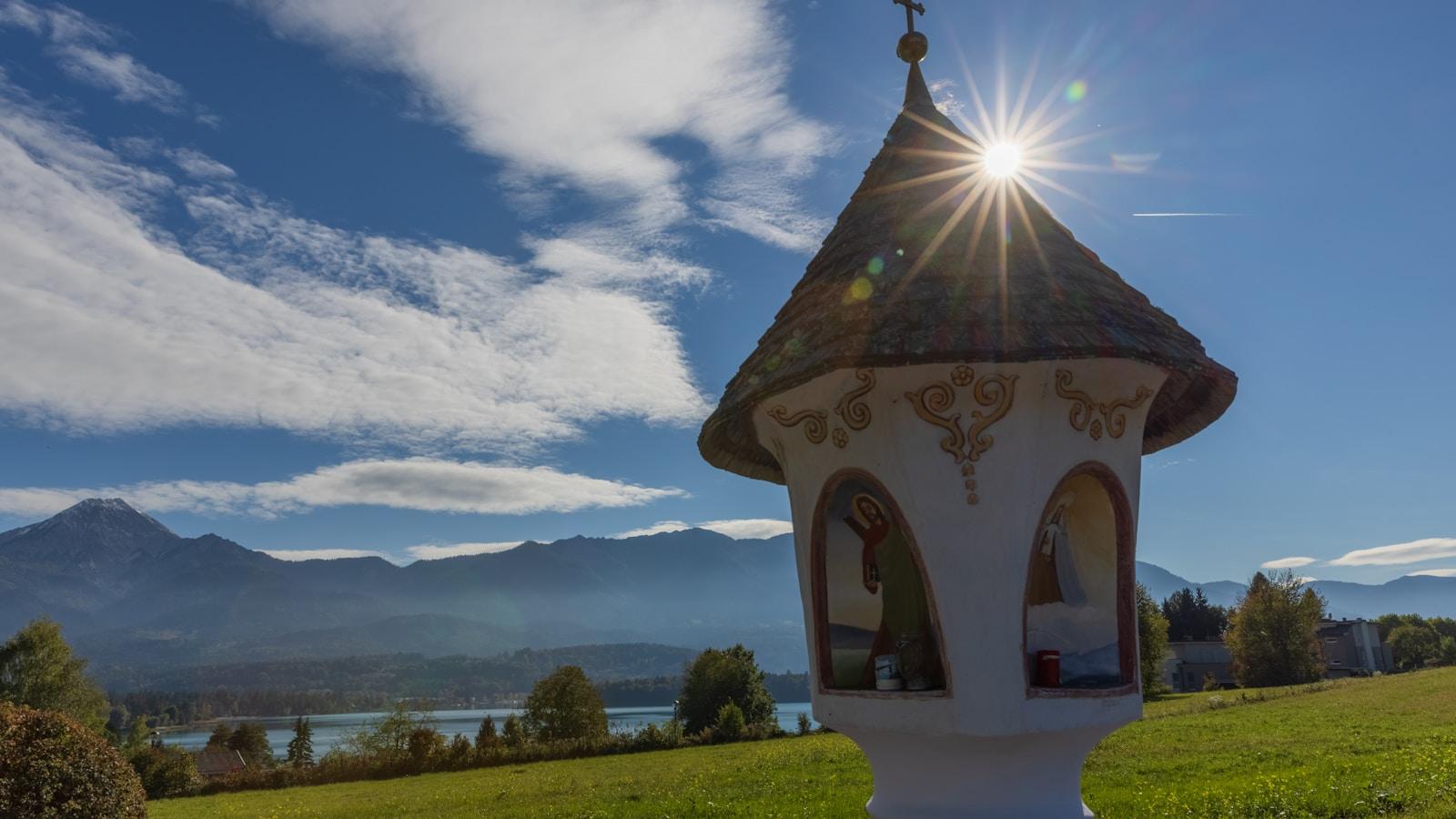 Pequeno oratório de pedra com telhado pontudo, diante de um lago e montanhas, sob o sol