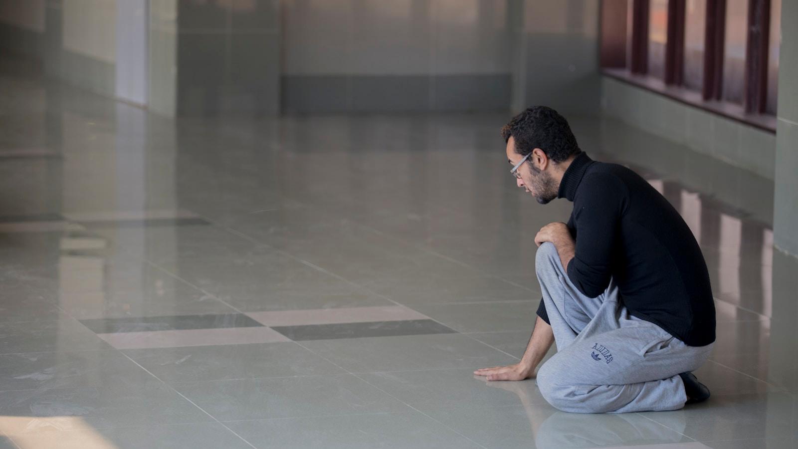 Homem ajoelhado no chão de um corredor amplo, com a mão apoiada no piso